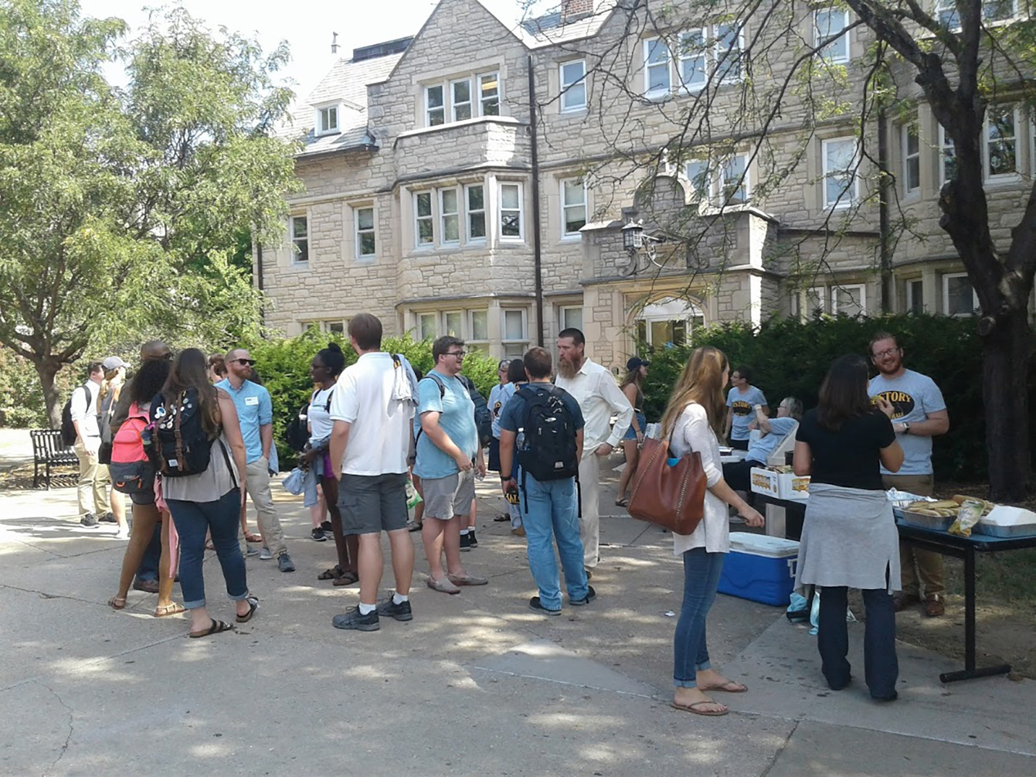 A group of people gather outdoors beside a stone building. Some stand near tables with food and drinks. The mood is casual and social.